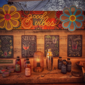 Wooden bar setup with 'good vibes' sign, flower decorations, and various bottles on a wooden surface.
